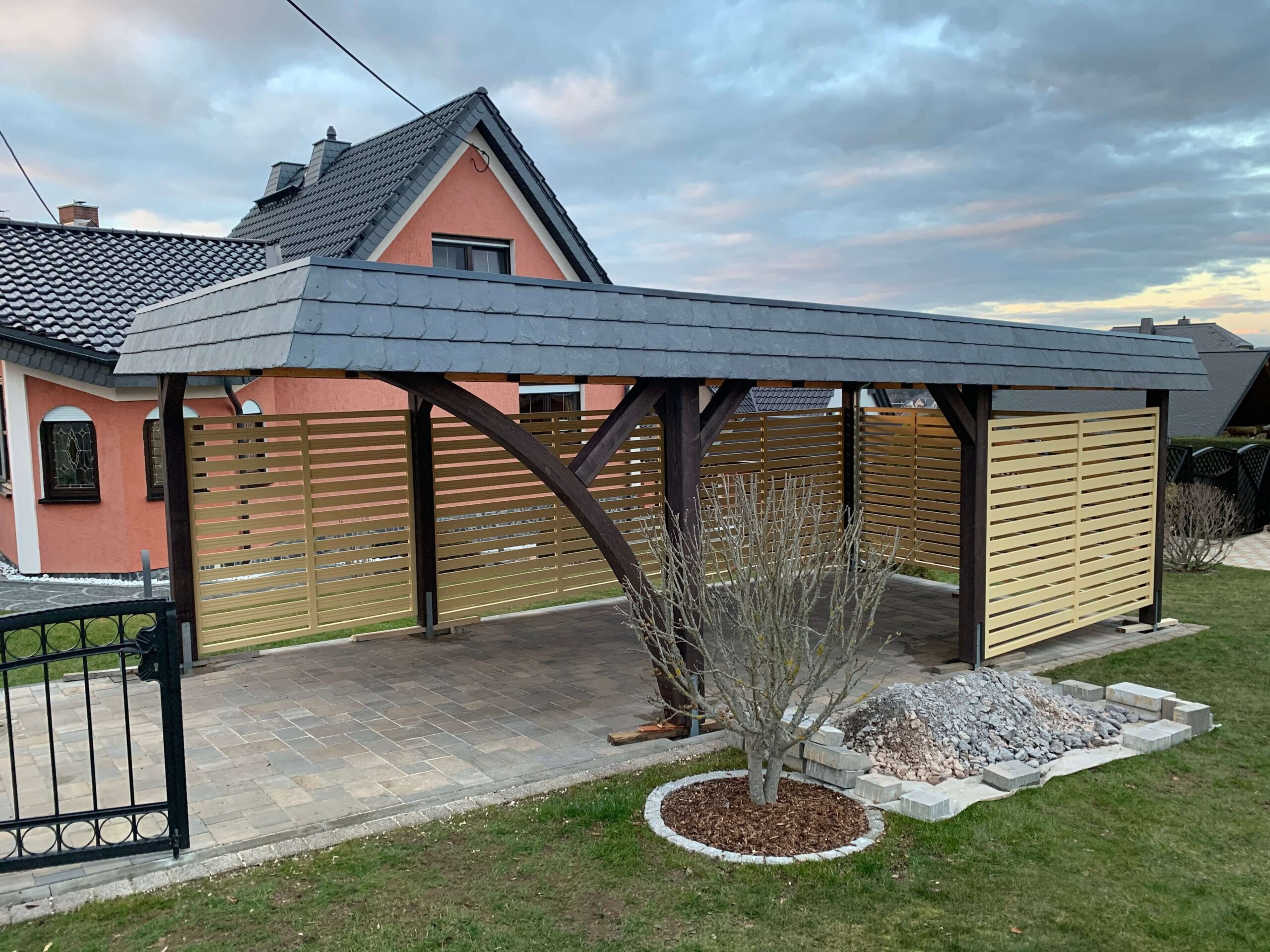 StyleLine Carport mit seitlichem Bogen, beiger Wandverkleidung und Walmdachblende in Schieferoptik. Die Kombination aus Braun und Schiefergrau wirkt warm und elegant zugleich.