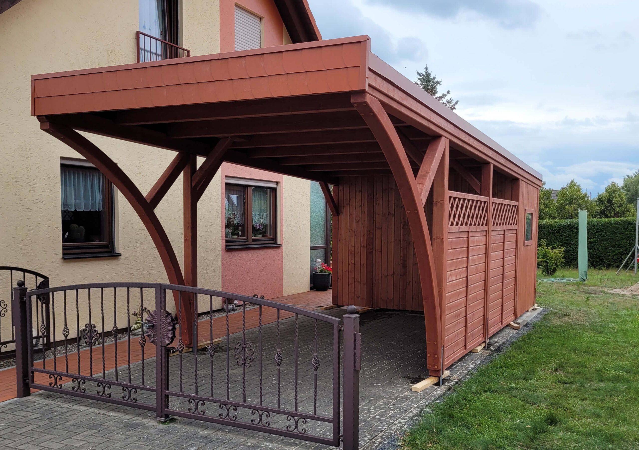 Einzelcarport mit Flachdach und zwei großen Bögen, gefertigt aus imprägniertem Mahagoniholz. Die Walmdachblende in Ziegelton unterstreicht den individuellen Charakter der Anlage.