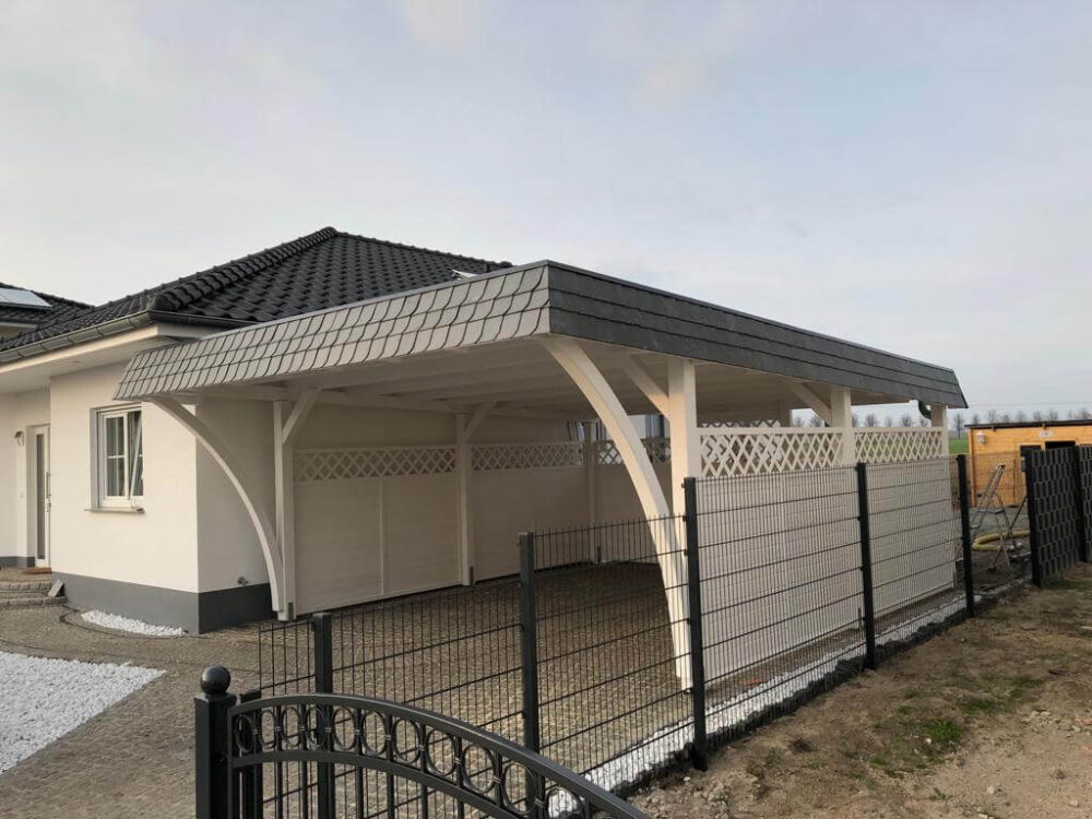 Weißer Doppelcarport mit Walmdachblende in Schieferoptik und eleganten Bögen. Die umlaufende Wandverkleidung sorgt für Struktur und geschützten Raum.