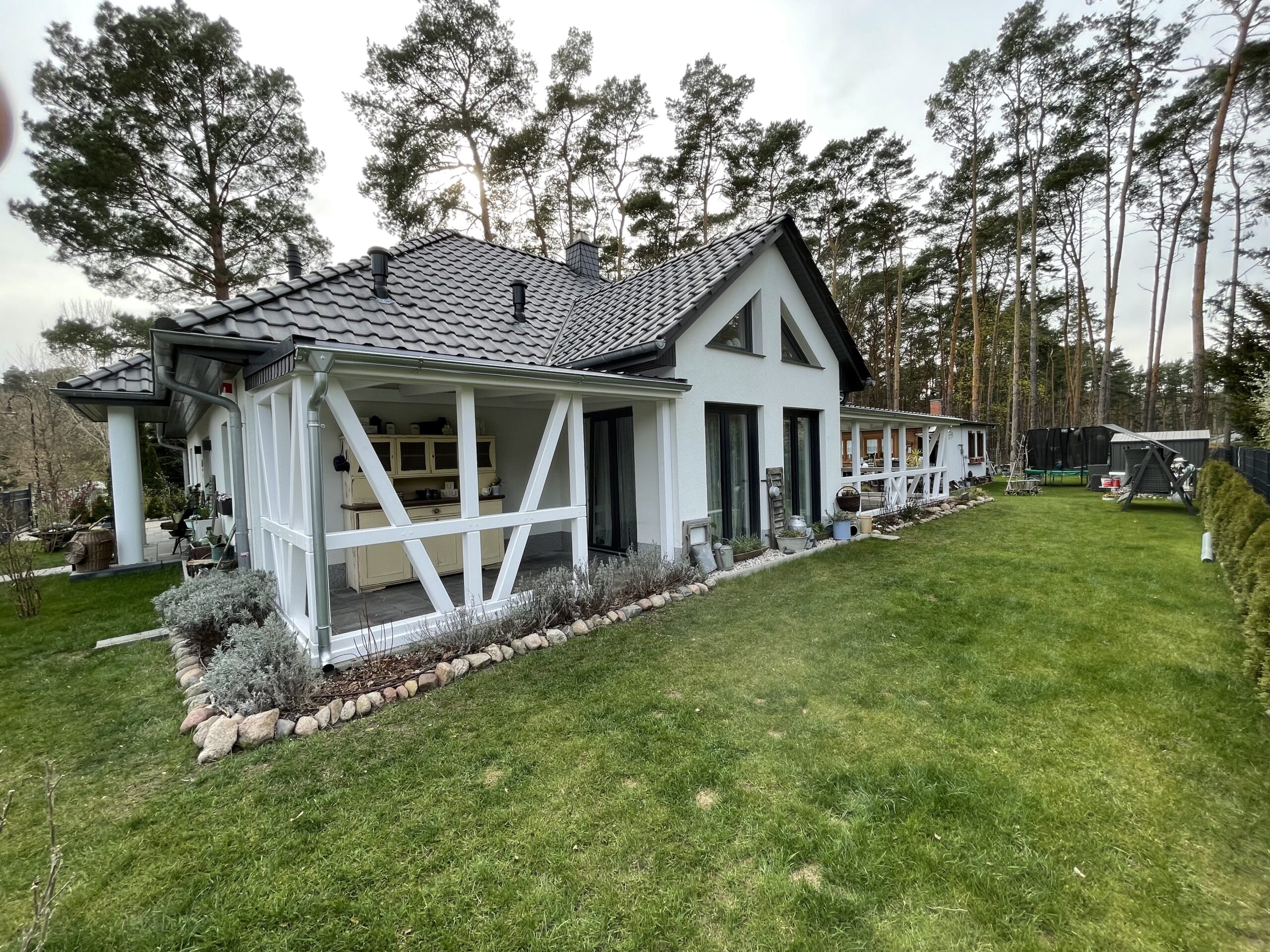 Großes L-förmiges Terrassendach in Fachwerkoptik, passend zum weißen Haus mit anthrazitfarbenem Dach. Holz in deckender Lackierung für edlen Kontrast.