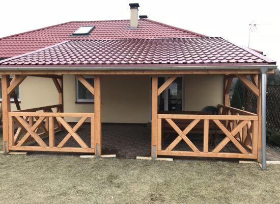 Frontansicht der Terrassenüberdachung mit Kreuzgeländer und Vivaldi-Dach in Dachziegeloptik. Harmonische Proportionen und massive Stützen aus Holz.